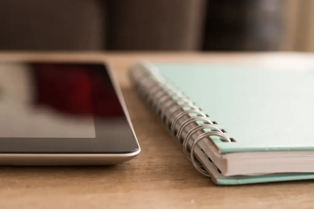 A tablet computer lying on a wooden desk next to a notebook and pen