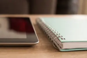 A tablet computer lying on a wooden desk next to a notebook and pen